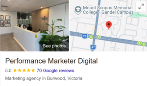 Top Left: A photograph showing the modern interior of an office reception desk area with a long white counter, warm lighting, indoor plants, and a person sitting behind the desk. A "See photos" overlay button is in the center. Top Right: A map snippet indicating the business's location with a red pin. Nearby landmarks shown include "Mount Scopus Memorial College - Gandel Campus" and roads labeled with the numbers 26 and 47. Bottom: The text elements of the profile, which include: The business name: Performance Marketer Digital A 5.0 star rating represented by five yellow stars, indicating 70 Google reviews. The business category and location listed below: Marketing agency in Burwood, Victoria. | Google Business Profile Optimisation Melbourne (2026 Guide)